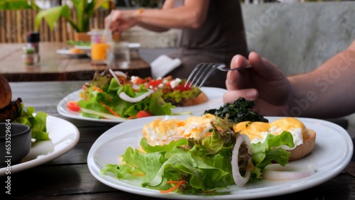 Man eating full meal as poached egg, salad and avocado toast in cafe. Close-up of man with fork pierces poached egg and eat tasty yolk. Order poached egg to eat healthy full meal. Full meal concept.