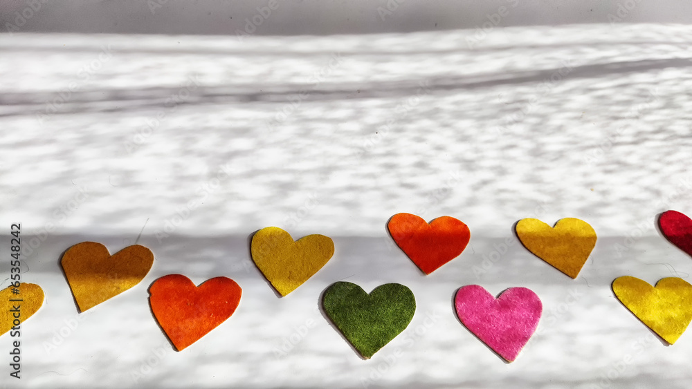 Fototapeta premium Multicolored hearts made of rough velvet paper on a white surface with stripes of shadow and light from the sun. Abstract background, texture, copy space
