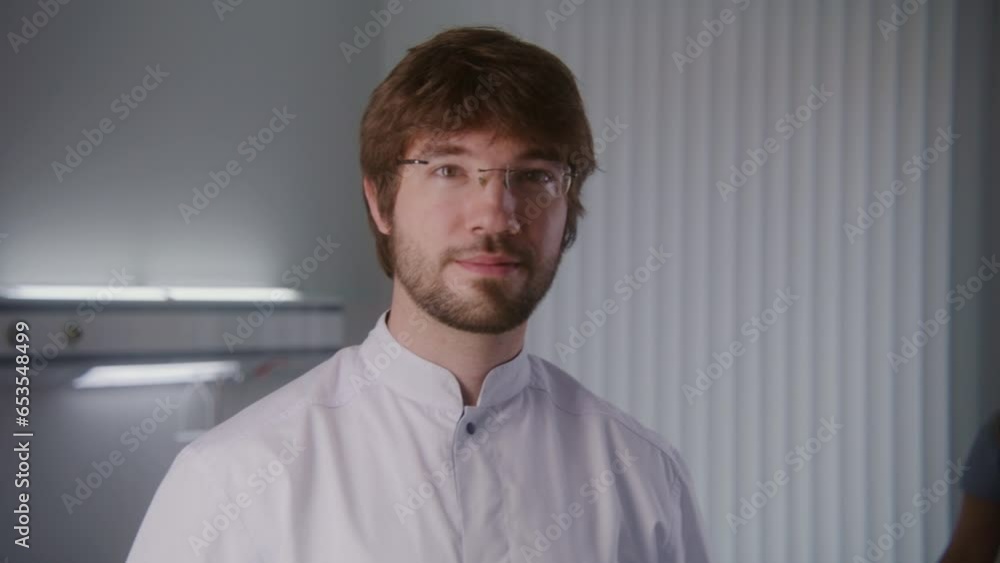 Adult medic in glasses stands in middle of hospital room. Professional healthcare specialist turns to camera and looks at it. Female health worker changes bedclothes on beds in the background. Adult medic in glasses stands in middle of hospital room. Professional healthcare specialist turns to camera and looks at it. Female health worker changes bedclothes on beds in the background.