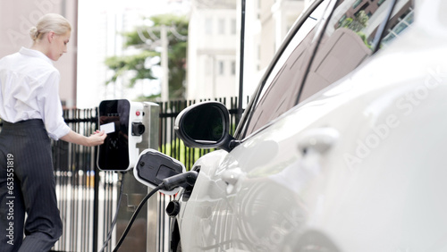 Progressive businesswoman install charger plug from charging station to his electric car before driving around city center. Eco friendly rechargeable car powered by sustainable and clean energy. photo