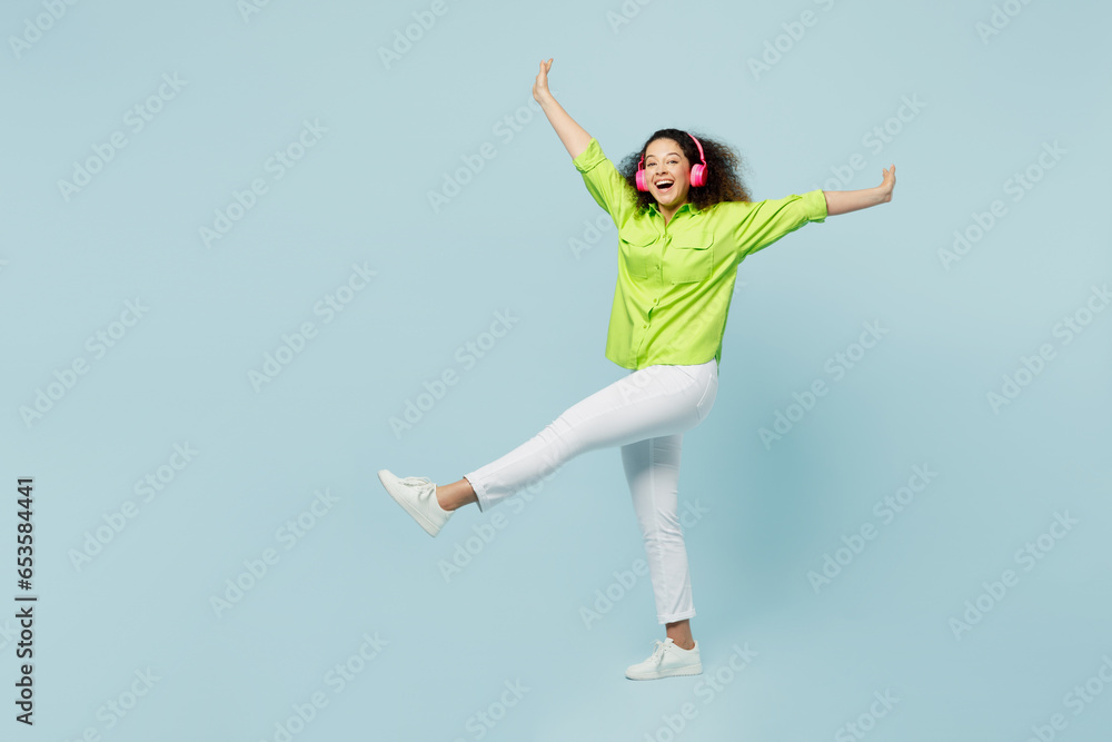 Full body young latin woman she wears green shirt casual clothes listen to music in headphones raise up hands leg dance isolated on plain pastel light blue cyan background studio. Lifestyle concept. Full body young latin woman she wears green shirt casual clothes listen to music in headphones raise up hands leg dance isolated on plain pastel light blue cyan background studio. Lifestyle concept.