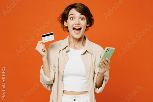 Young happy caucasian woman she wear beige shirt casual clothes using mobile cell phone hold credit bank card doing online shopping order delivery booking tour isolated on plain orange red background.