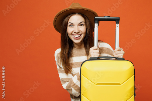 Traveler happy cheerful woman wear brown sweater hat hold suitcase bag isolated on plain orange red background. Tourist travel abroad in free spare time rest getaway. Air flight trip journey concept.
