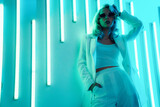 Young beautiful hipster woman in street style fashion concept. Hot model wearing white suit and sunglasses. Female posing in blue neon light in studio interior