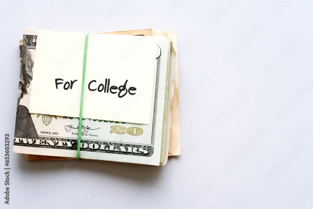 Dollars cash money in rubber band with text written note FOR COLLEGE , on copy space background - concept of financial planning to save money for purpose of education Dollars cash money in rubber band with text written note FOR COLLEGE , on copy space background - concept of financial planning to save money for purpose of education