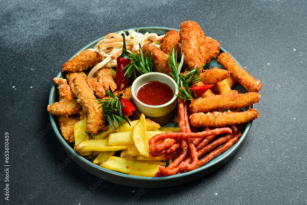 Fast food. Plate with snacks: nuggets, sausages, French fries, cheese sticks. On a black stone background. Free space for text. Fast food. Plate with snacks: nuggets, sausages, French fries, cheese sticks. On a black stone background. Free space for text.