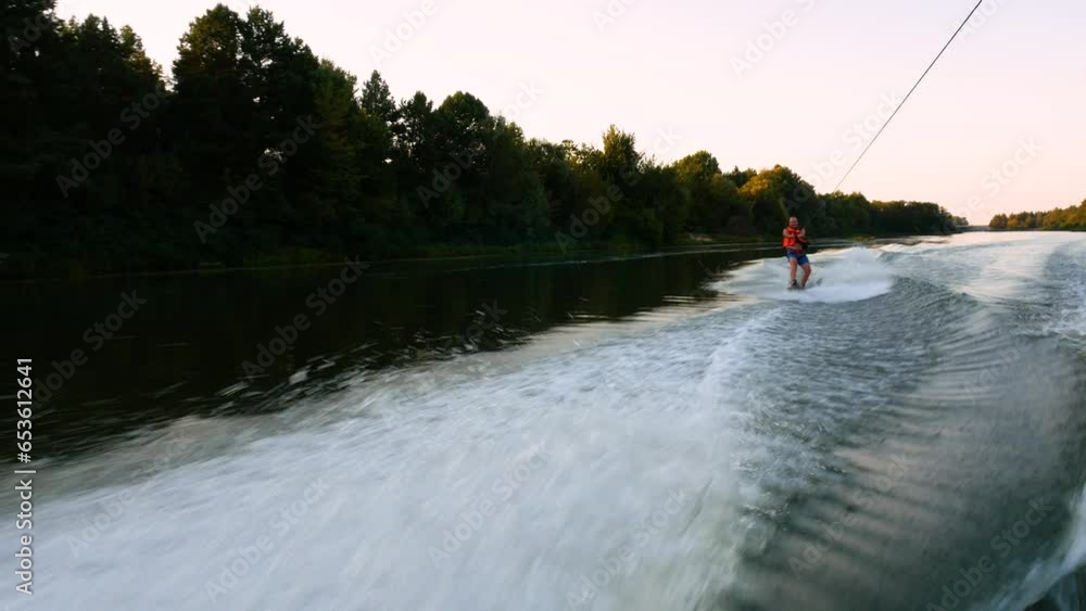 Man water skiing on a lake. River sports. A man rides a board on the river. Waterboarding on the river. Man in red life vest waterboarding. The wave behind the boat. Man water skiing on a lake. River sports. A man rides a board on the river. Waterboarding on the river. Man in red life vest waterboarding. The wave behind the boat.
