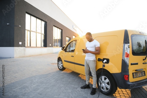 Casual african man with smartphone near electric car waiting for the finish of the battery charging process.