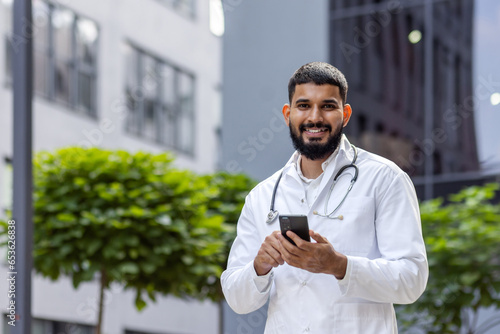 Portrait of a young Muslim male doctor standing smiling near the clinic and holding a phone in his hands. Looking and smiling at the camera
