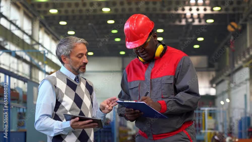 Black Worker in Protective Workwear Taking Notes on Clipboard and Production Supervisor Signing a Document in a Factory. Caucasian Production Manager Giving Instructions To Black Industrial Worker.