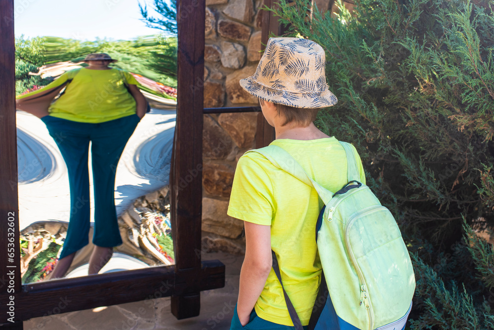 the boy stands in front of a distorting mirror. a child looks at his ...