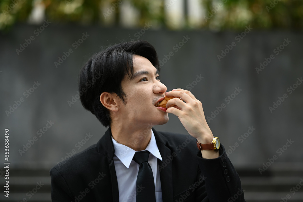 Happy hangry businessman having lunch outdoors, eating sandwich while sitting on stairs outside office building Happy hangry businessman having lunch outdoors, eating sandwich while sitting on stairs outside office building