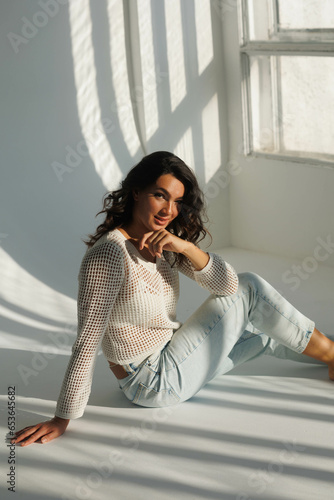 Beautiful brunette woman in white net top and blue jeans, sitting. Girl smile, happy. Portrait of young pretty woman. Complicated sunlight with shadows. White background. Light from window, portrait.