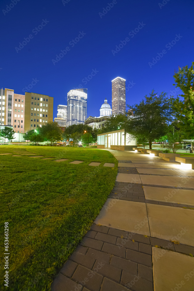 Illuminated pathway concrete sidewalk at park square with downtown Houston skyscraper, capitol tower in background during evening blue hour