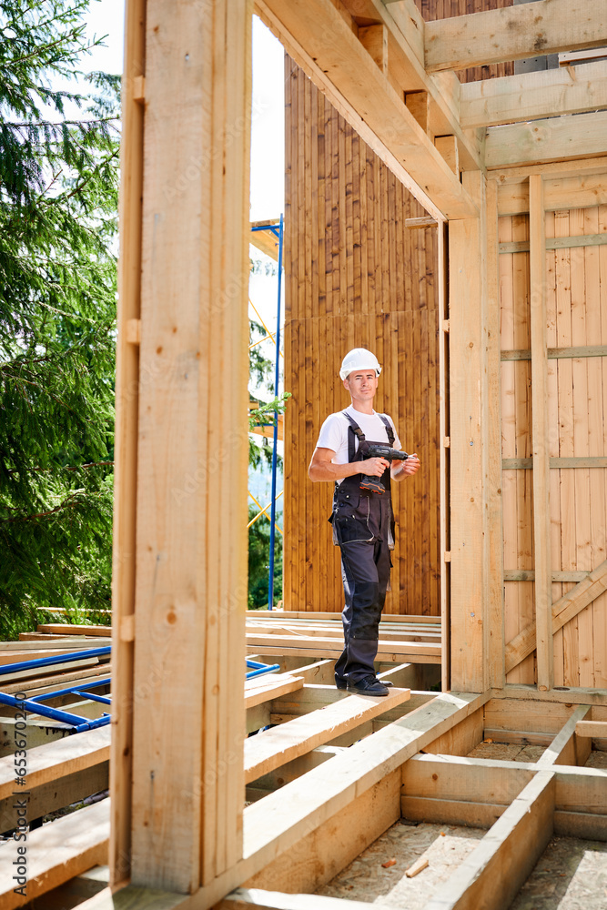 Portrait of carpenter constructing wooden framed house. Man worker cladding facade of house, using screwdriver. Concept of modern eco-friendly construction. Portrait of carpenter constructing wooden framed house. Man worker cladding facade of house, using screwdriver. Concept of modern eco-friendly construction.