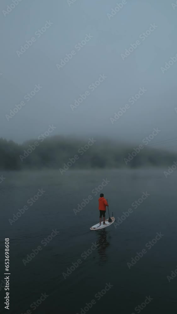 Aerial vertical view of man on sup board paddling doing water sport activity on mountain lake morning misty fog floating over lake. Healthy lifestyle outdoor activity Aerial vertical view of man on sup board paddling doing water sport activity on mountain lake morning misty fog floating over lake. Healthy lifestyle outdoor activity