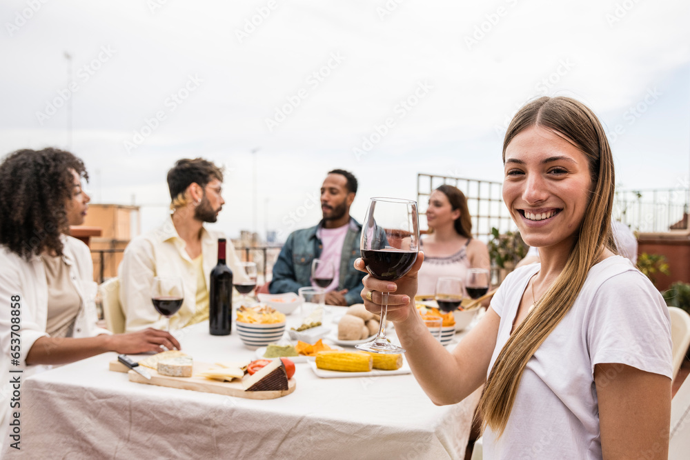 Young cheerful lady looking at camera with a glass of wine in a rooftop dinner party. Joyful beautiful woman toasting and staring at camera at outdoors terrace. Young cheerful lady looking at camera with a glass of wine in a rooftop dinner party. Joyful beautiful woman toasting and staring at camera at outdoors terrace.