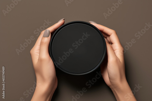 Female hands holding cosmetics black jar mockup on brown background, top view. Moisturizing and nourishing, beauty cosmetic product industry, skin care concept