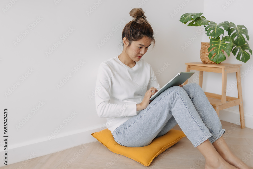 Happy asian Thai woman bun hair, sitting on floor, using tablet, working at apartment in winter day.