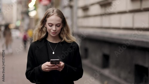 Pretty young blond woman hold smartphone scrolling social media texting browsing online while walking down street in cold city centre Positive female relax enjoying great autumn day outdoors 