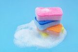 Stack of many colorful sponges in soap foam on blue background. Cleaning and washing concept. 