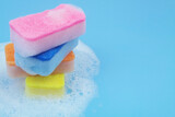 Stack of many colorful sponges in soap foam on blue background. Cleaning and washing concept. Copy space for text.