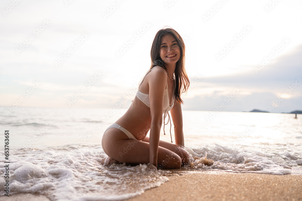 Beautiful caucasian woman in white bikini on tropical beach. Portrait of dark skinned woman smiling at sea. Brunette tanned girl in swimwear enjoying and walking on beach.