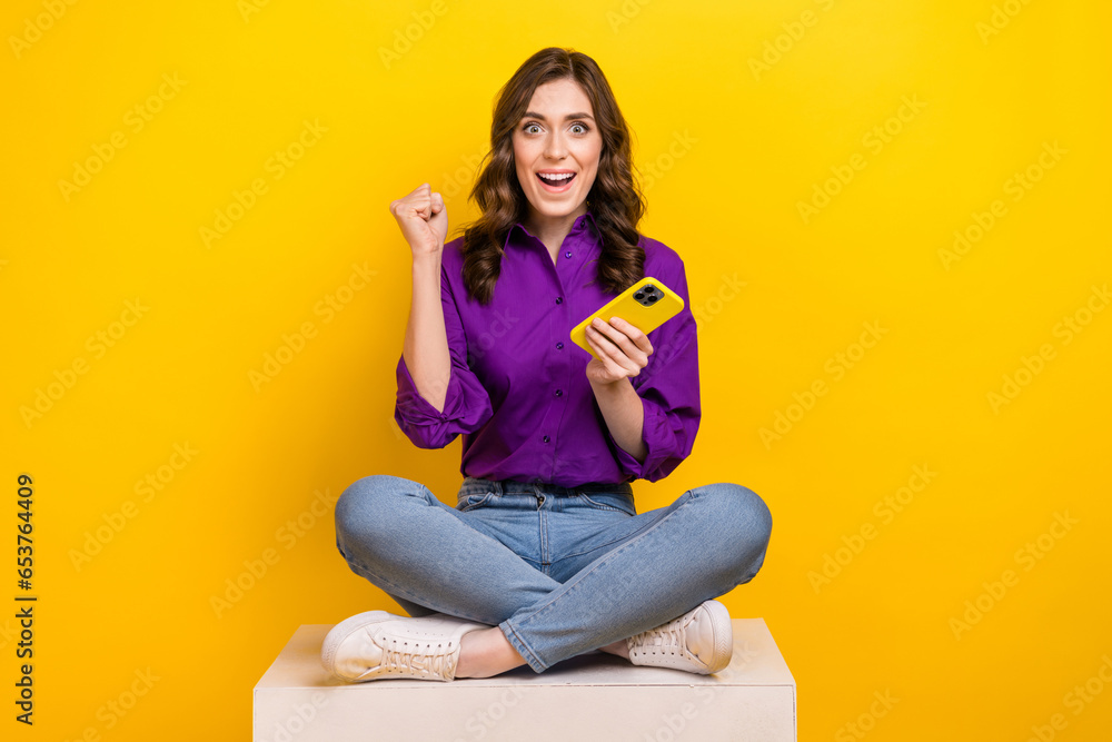 Full body photo of delighted crazy lady sit podium cube hold smart phone raise fists isolated on yellow color background Full body photo of delighted crazy lady sit podium cube hold smart phone raise fists isolated on yellow color background