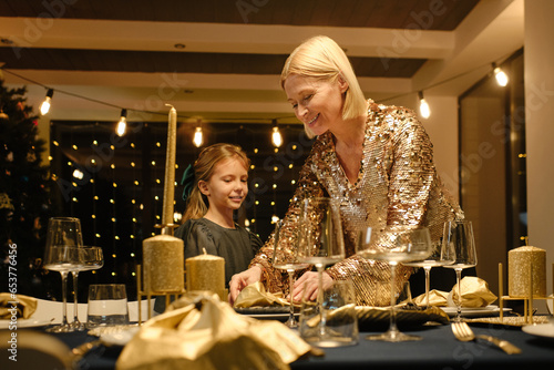 Portrait of mature woman and girl wearing fancy outfits preparing to celebrate Christmas finishing dining table setting photo