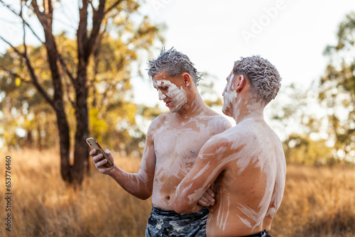 Teen aboriginal cousins in traditional ochre body paint using mobile phone social media photo