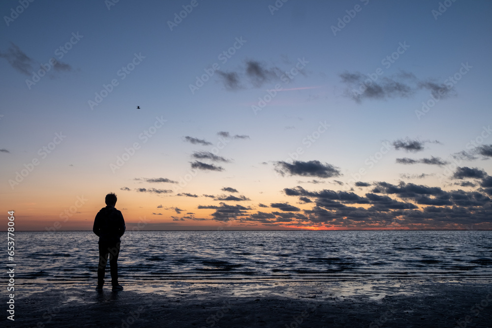 Witness the serene beauty of a moment in time as a young man stands on the tranquil beach, his silhouette framed against the breathtaking backdrop of a dramatic and colorful after-sunset sky. With the