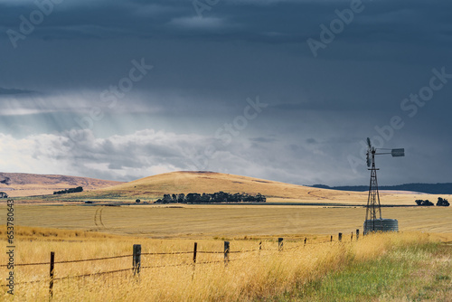 A windmill on the roadside of dry rural farmland with dark clouds and rain falling in the distance photo