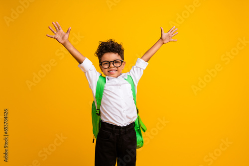 Portrait of funky overjoyed schoolkid toothy smile carry backpack raise hands empty space isolated on yellow color background