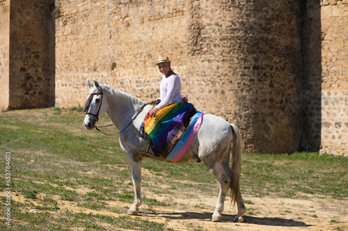 Young non-binary latin person, riding a white horse with the gay pride flag. In the background a medieval castle. Concept of diversity, homosexuality and human rights. © @skuder_photographer