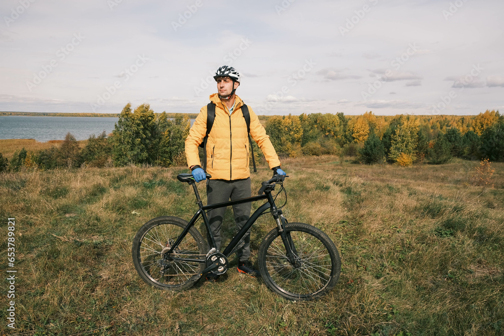 man in a protective helmet and a backpack stands next to a mountain bike in autumn against the background of a forest and a lake.An active lifestyle.Cycling man in a protective helmet and a backpack stands next to a mountain bike in autumn against the background of a forest and a lake.An active lifestyle.Cycling