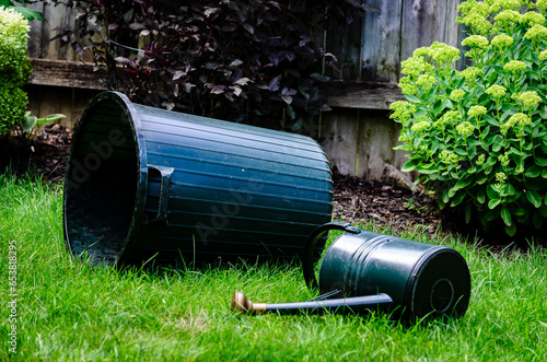 Empty garbage pail and watering can on their sides in a fenced backyard