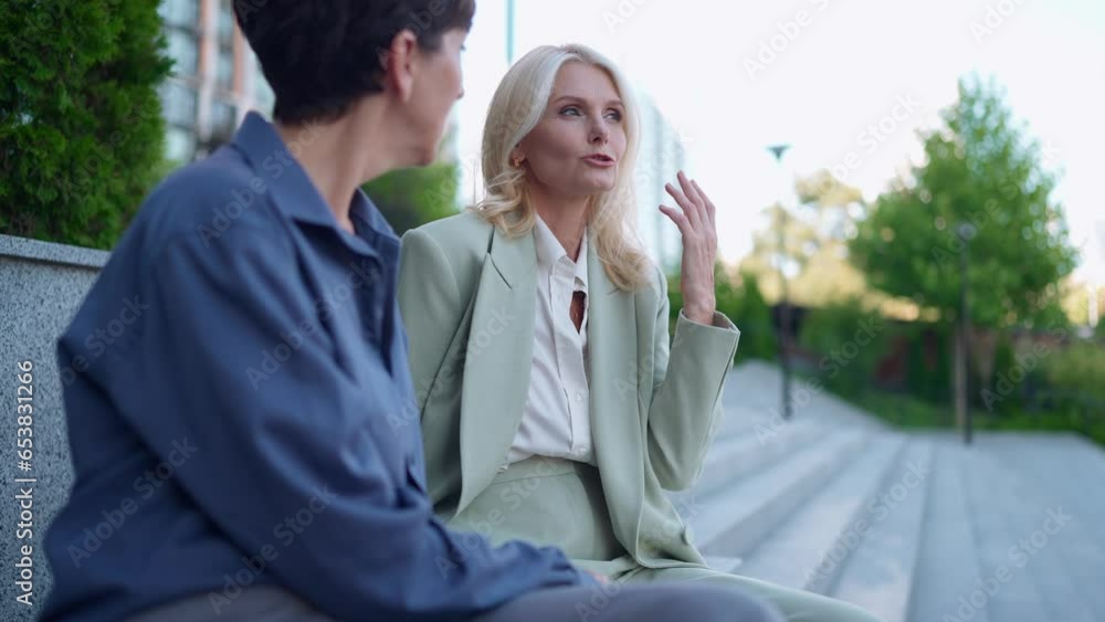 Pensive mature businesswomen in suits talking sitting on the steps outside Pensive mature businesswomen in suits talking sitting on the steps outside
