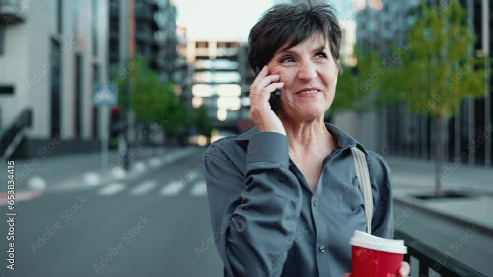 Smiling brunette mature businesswoman in shirt talking on cell phone and walking with cup of coffee outdoors Smiling brunette mature businesswoman in shirt talking on cell phone and walking with cup of coffee outdoors