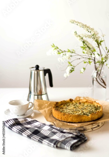 There is a quiche pie on parchment paper, next to it there is a coffee pot and a white cup and saucer. There is a vase of white flowers on the table and a black and white checkered napkin