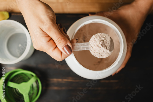 Female hand holds measuring spoon with portion of whey protein powder above plastic jar on wooden table with shaker, banana and apple fruit. Process of making protein drink, top view
