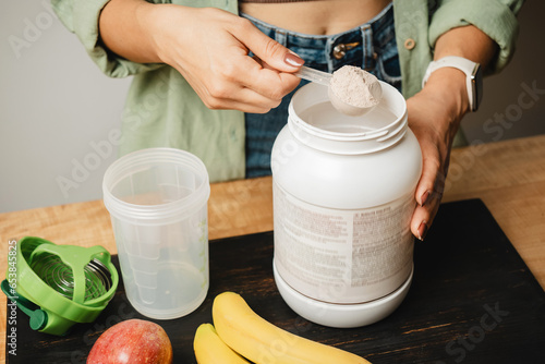 Woman in a jeans and shirt holds a measuring spoon with portion of whey protein powder above plastic jar on wooden table with shaker, banana and apple fruit. Process of making protein cocktail drink