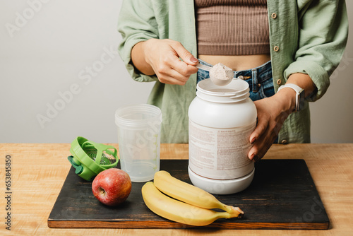 Fotografía Woman in a jeans and shirt holds a measuring spoon with portion of whey protein powder above plastic jar on wooden table with shaker, banana and apple fruit
