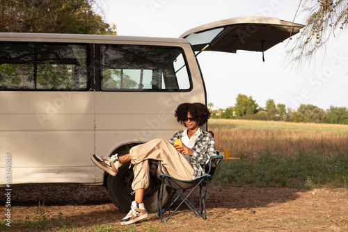 young african black woman using her smartphone in the nature in a chair beside her camper van during a road trip at autumn