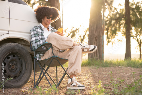 Young African black woman in the nature using her smartphone during a road trip in her camper van at autumn