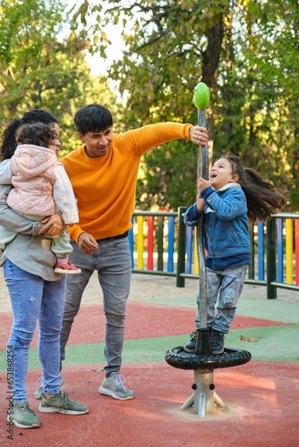 Happy Hispanic parents playing with her children at the synchro spinner in a park. Latin family.