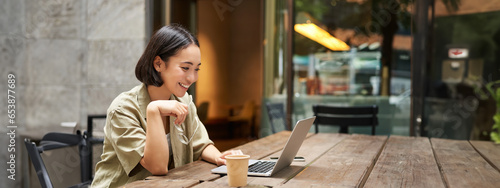 Portrait of asian woman looking at laptop, video chat, talking with someone via computer camera, sitting in cafe and drinking coffee, online meeting