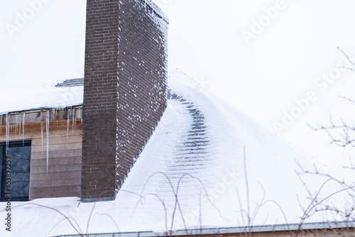 A portrait of a roof of a house full of white snow, except for a trail which melted due to bad isolation. There is a big brick chimney and the roof got covered after a snowstorm or blizzard in winter. photo