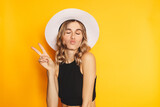 Portrait of a happy cheerful girl in white hat and black top showing peace gesture with two fingers, blow kiss, closed eyes isolated over yellow background. Victory peace gesture blonde hair girl.