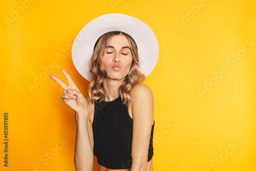 Portrait of a happy cheerful girl in white hat and black top showing peace gesture with two fingers, blow kiss, closed eyes isolated over yellow background. Victory peace gesture blonde hair girl.