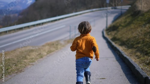 From behind, a spirited child in a yellow pullover, jeans, and boots sprints with delight through the autumn scenery. The lively run embodies the thrill of the crisp fall air